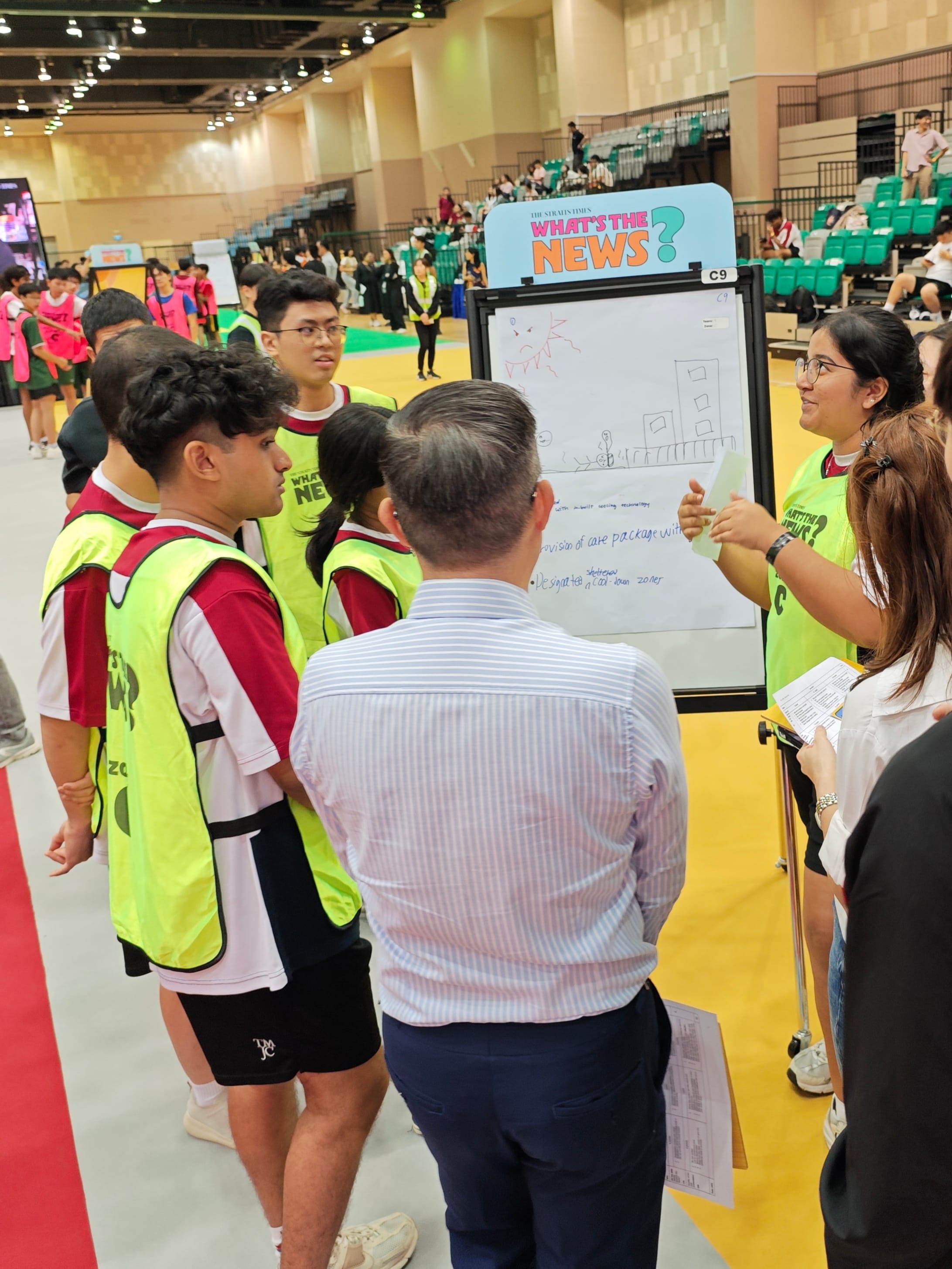 A small group of students in yellow vests presenting ideas on a whiteboard to a judge at a large indoor venue.